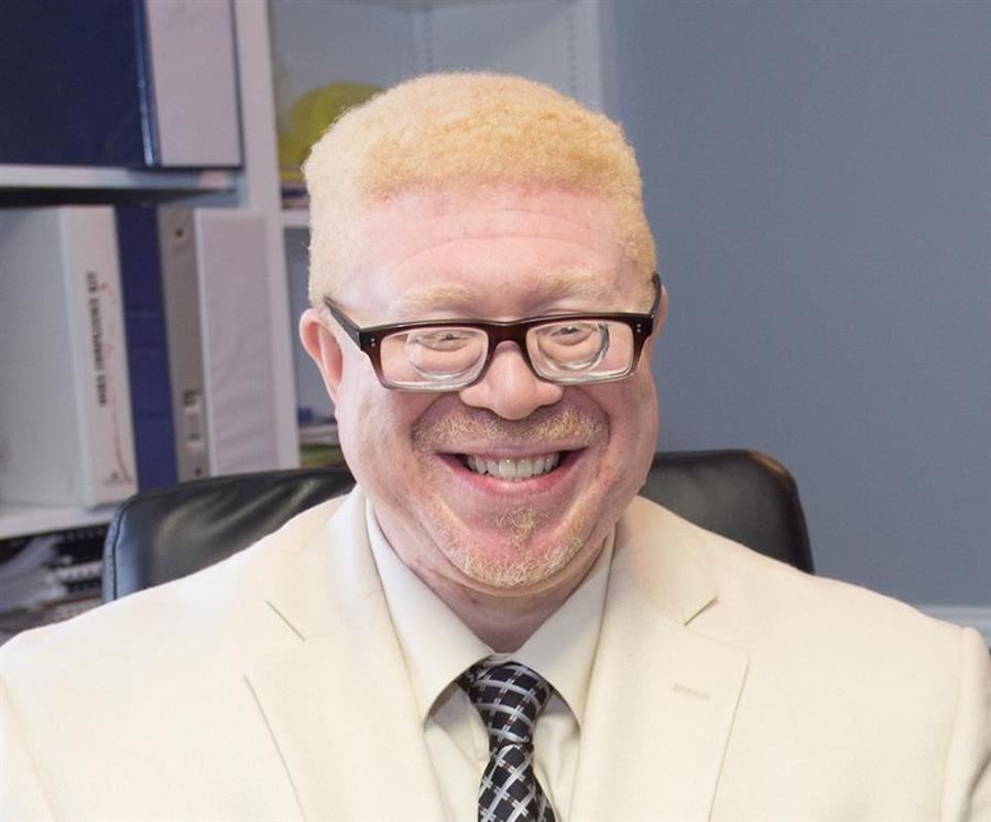 Isaac Beavers, wearing a tan suit and glasses, smiles to camera.